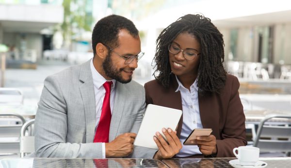 Cheerful coworkers looking at each other gadgets screen. Business man and woman sitting in coffee shop, showing tablet and smartphone screens to each other. Business and modern technology concept