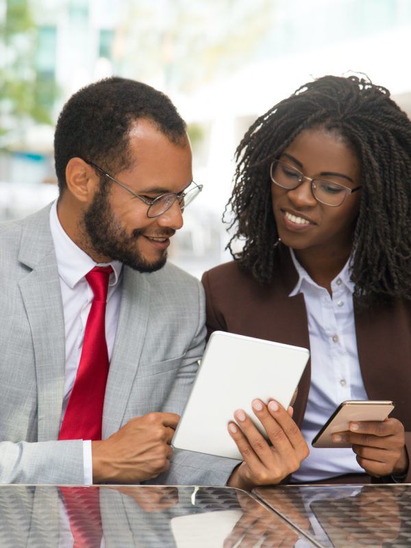 Cheerful coworkers looking at each other gadgets screen. Business man and woman sitting in coffee shop, showing tablet and smartphone screens to each other. Business and modern technology concept